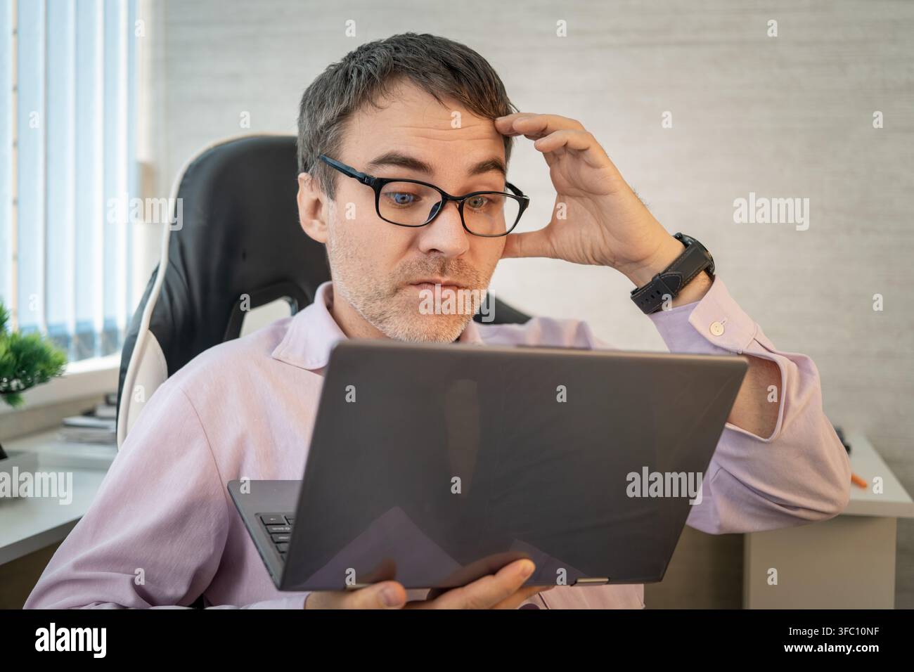 Confused office worker touching his forehead while looking at his laptop screen with a skeptical expression. Misunderstanding, difficult task, or read Stock Photo