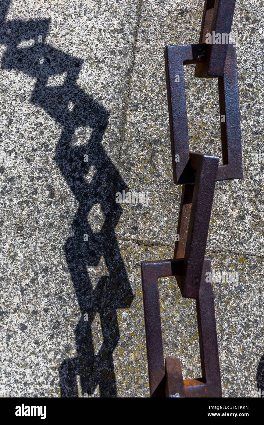A heavy iron chain casts a sharp, geometric shadow onto a textured stone surface outdoors in Spain. Stock Photo