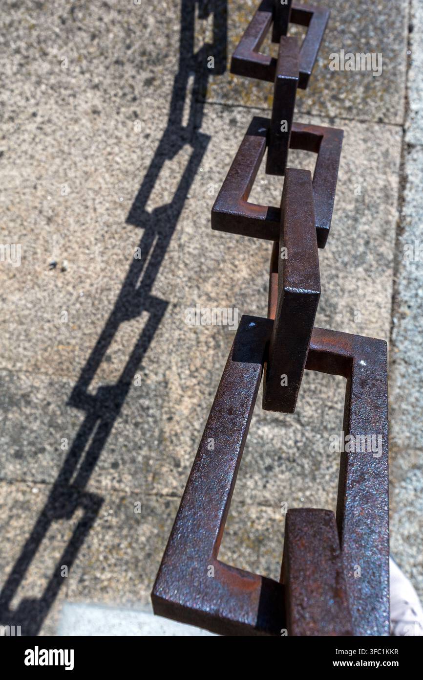 A heavy iron chain casts a sharp, geometric shadow onto a textured stone surface outdoors in Spain. Stock Photo