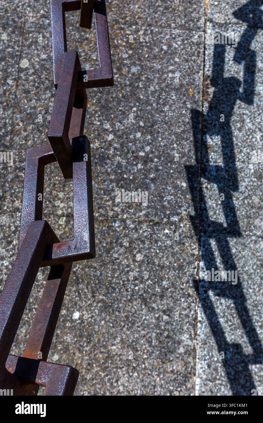 A heavy iron chain casts a sharp, geometric shadow onto a textured stone surface outdoors in Spain. Stock Photo