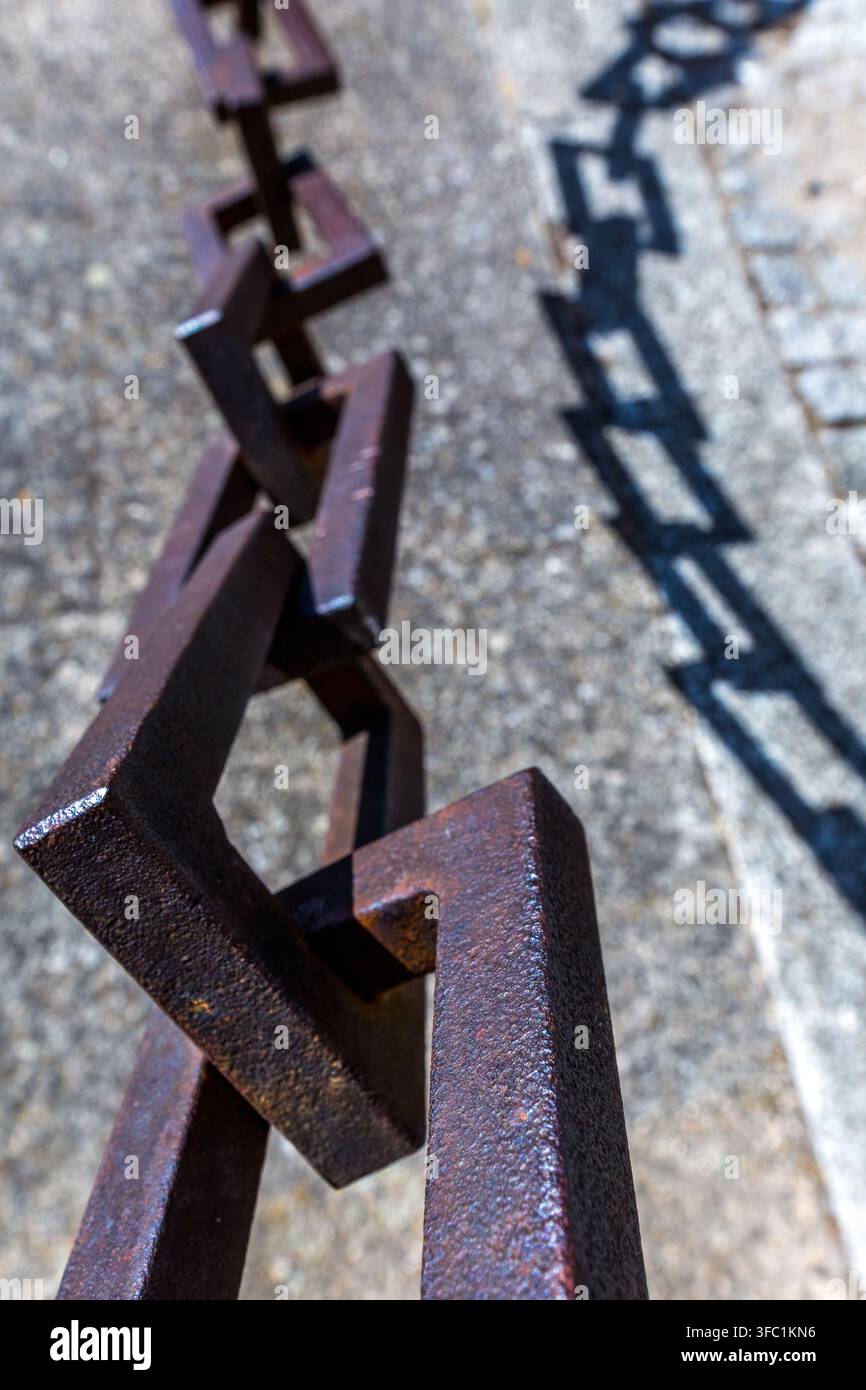 A heavy iron chain casts a sharp, geometric shadow onto a textured stone surface outdoors in Spain. Stock Photo