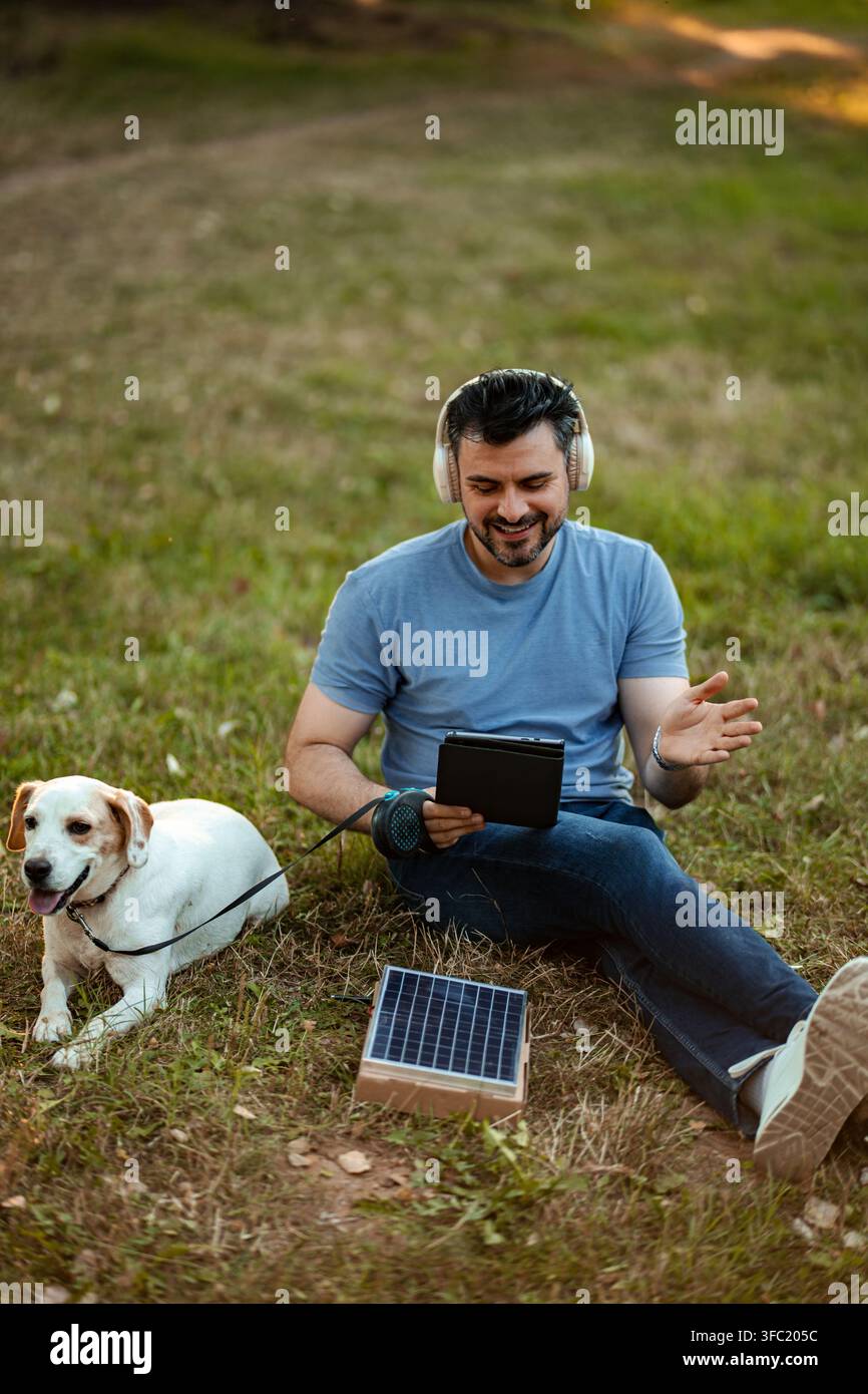 Man and dog relaxing in park, using solar power to charge tablet Stock Photo