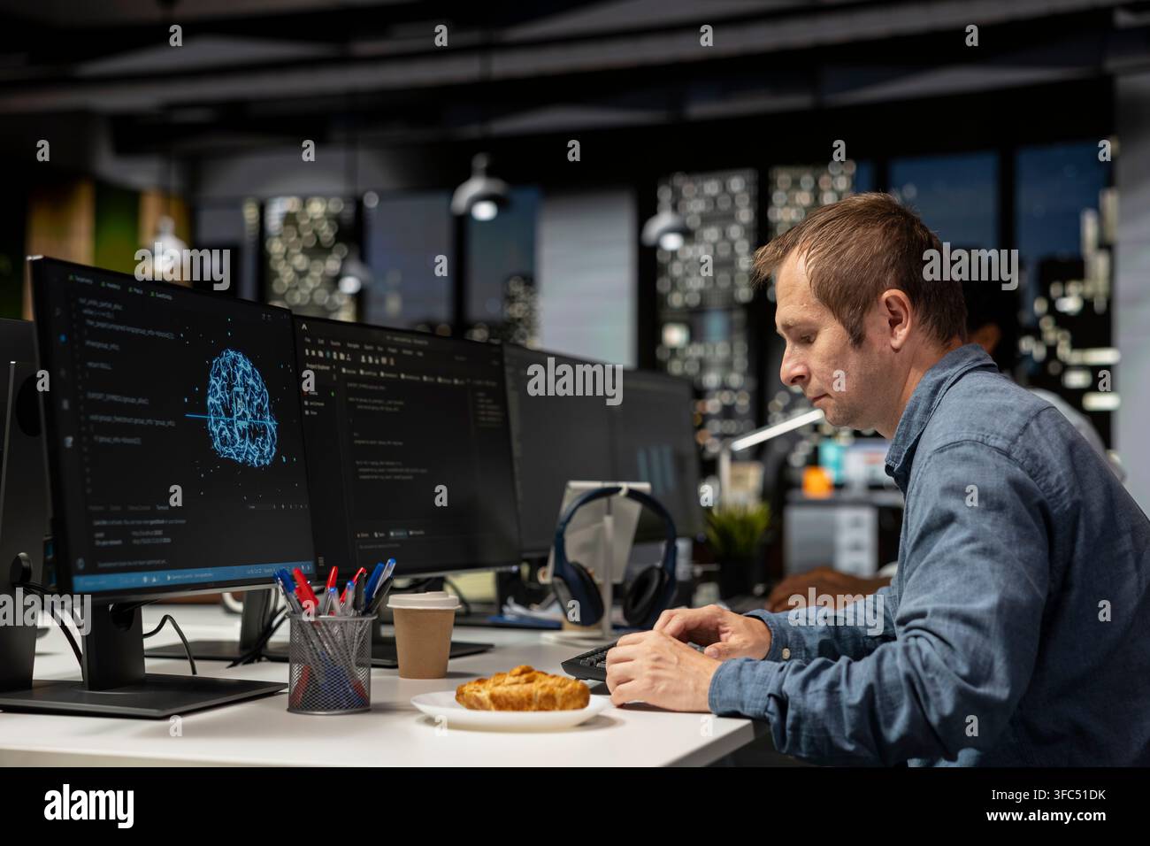Engineer utilizing AI on computer to optimize performance while eating croissant. Worker in tech company office taking snack break while training machine learning models to perform automatic updates Stock Photo
