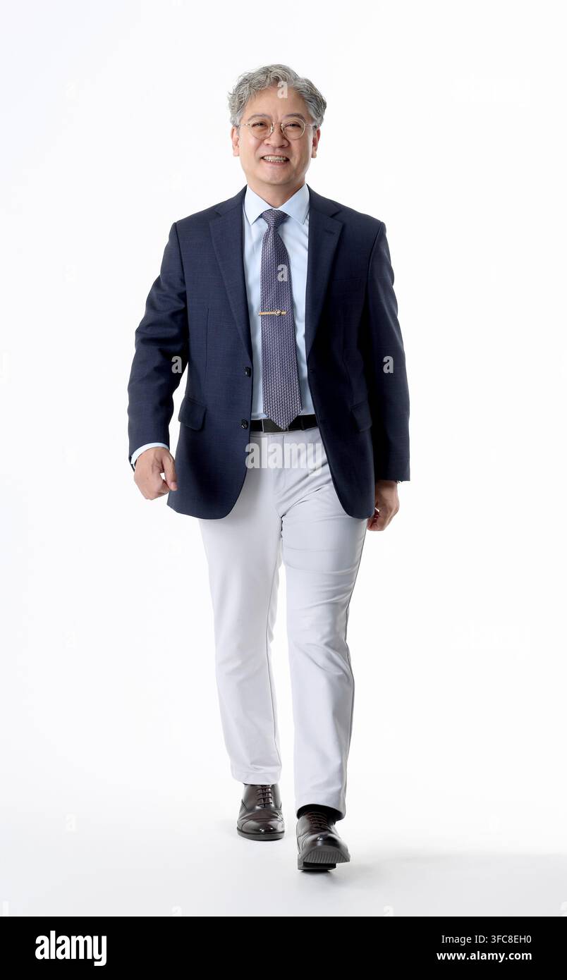 Confident senior Korean executive walking forward in formal business attire against a white background. Stock Photo