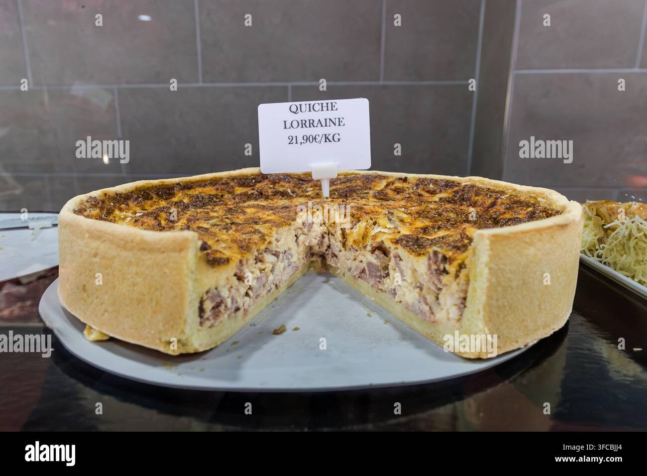 A sliced Quiche Lorraine sits with a price sign reading "21.90€/kg" in a display case in Nancy, Grand Est, France. The golden-brown bacon-and-egg filling is clearly visible. Stock Photo