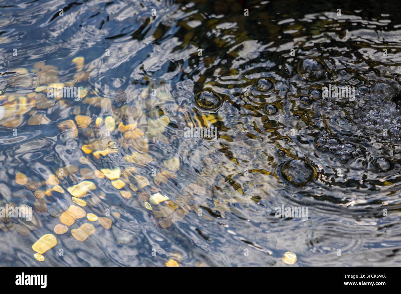 Coins rest underwater in a fountain. Ripples and bubbles shape the surface. Light reflections create soft patterns on the metal. Stock Photo