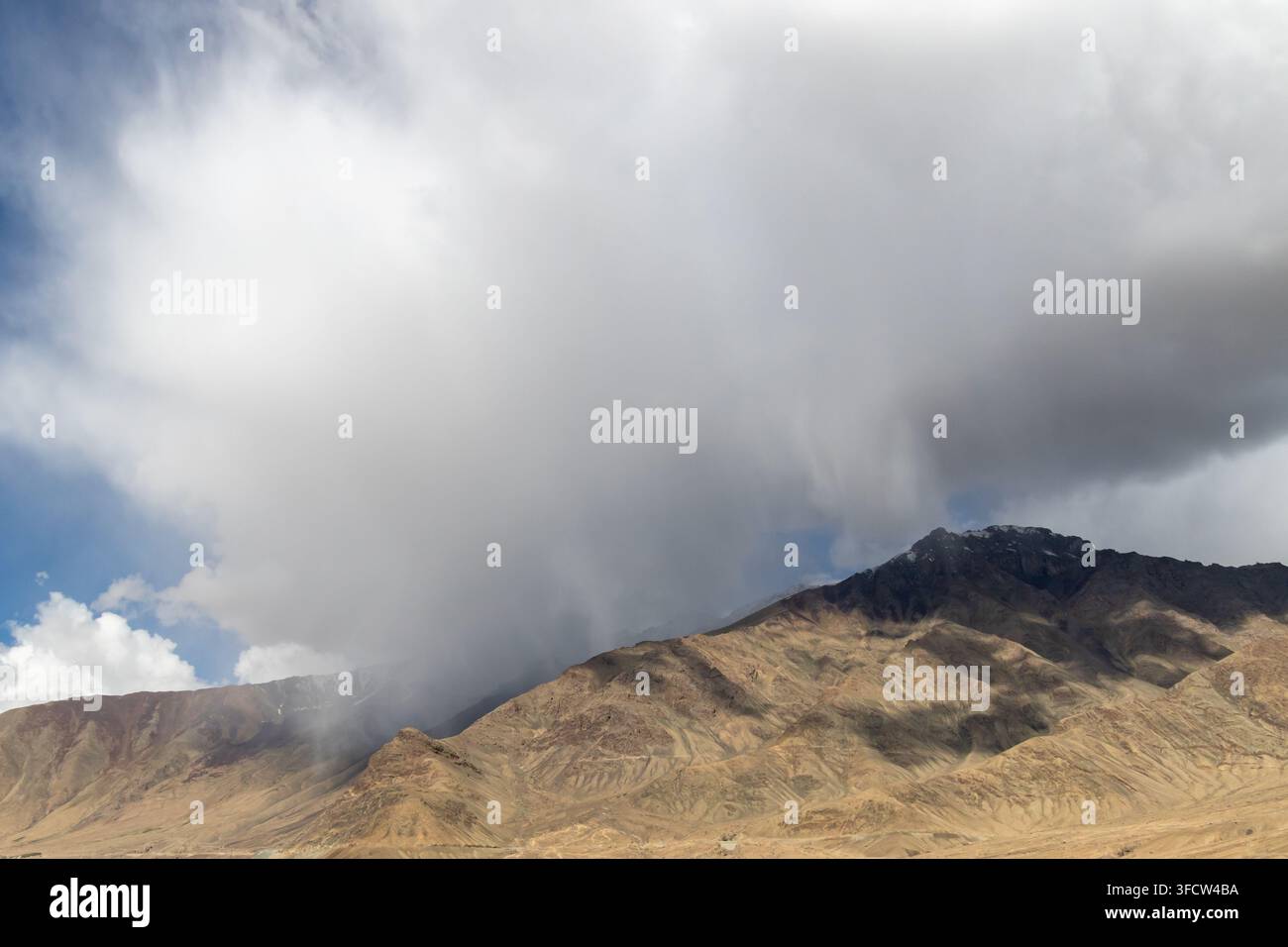 Dramatic storm clouds sweep over dry brown mountain ridges in a rugged highland landscape, with patches of blue sky and sunlight on the slopes. Stock Photo