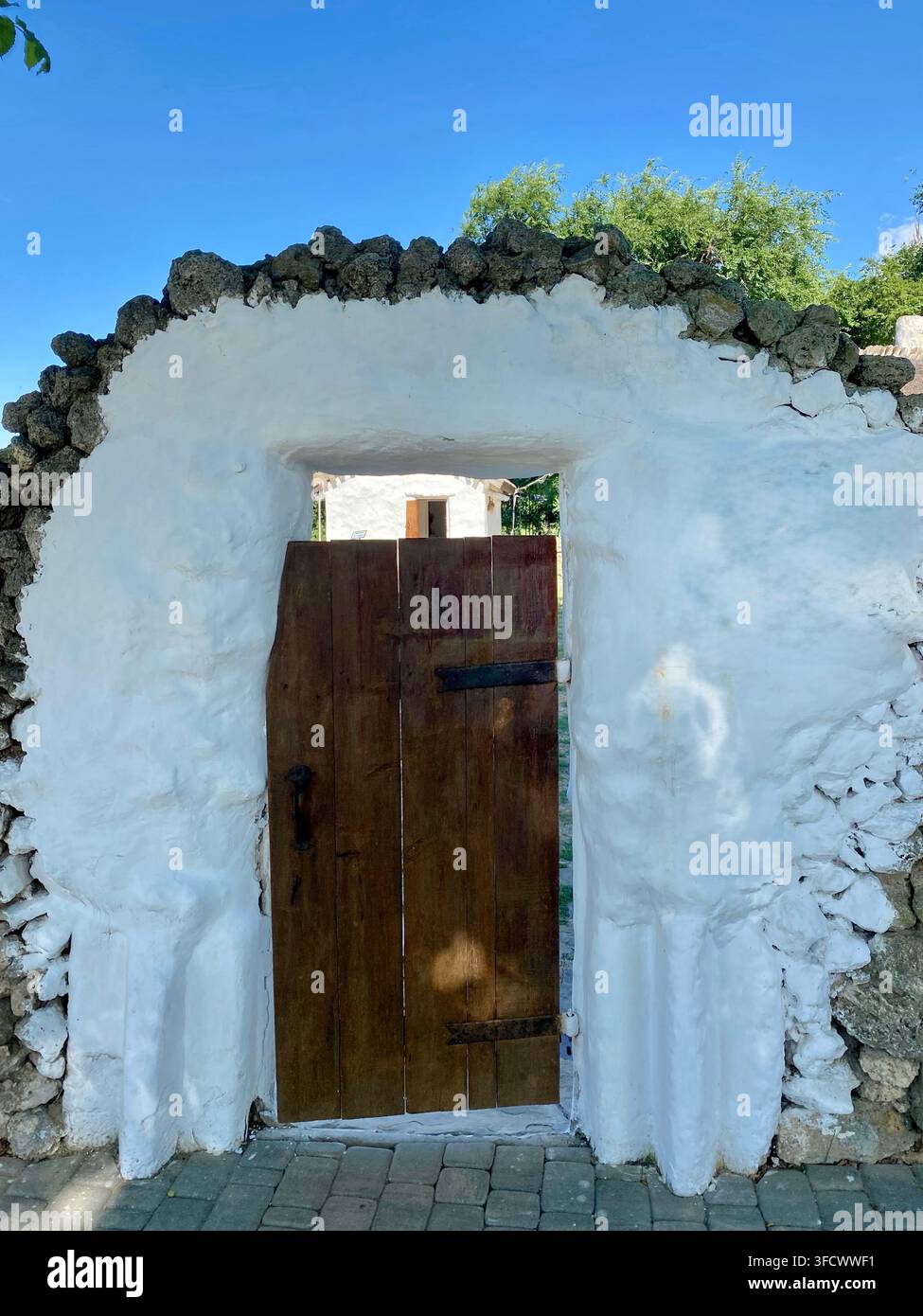 Rustic wooden gate framed by whitewashed stone wall under blue sky. Stock Photo