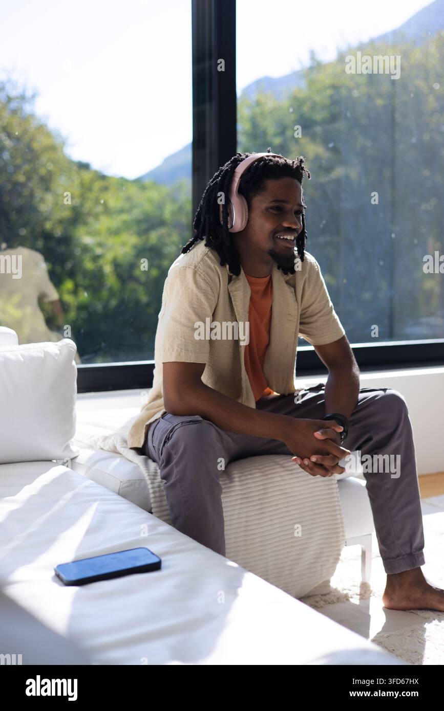 African American man sitting on ottoman at home, wearing orange tee, pink headset, phone on couch Stock Photo