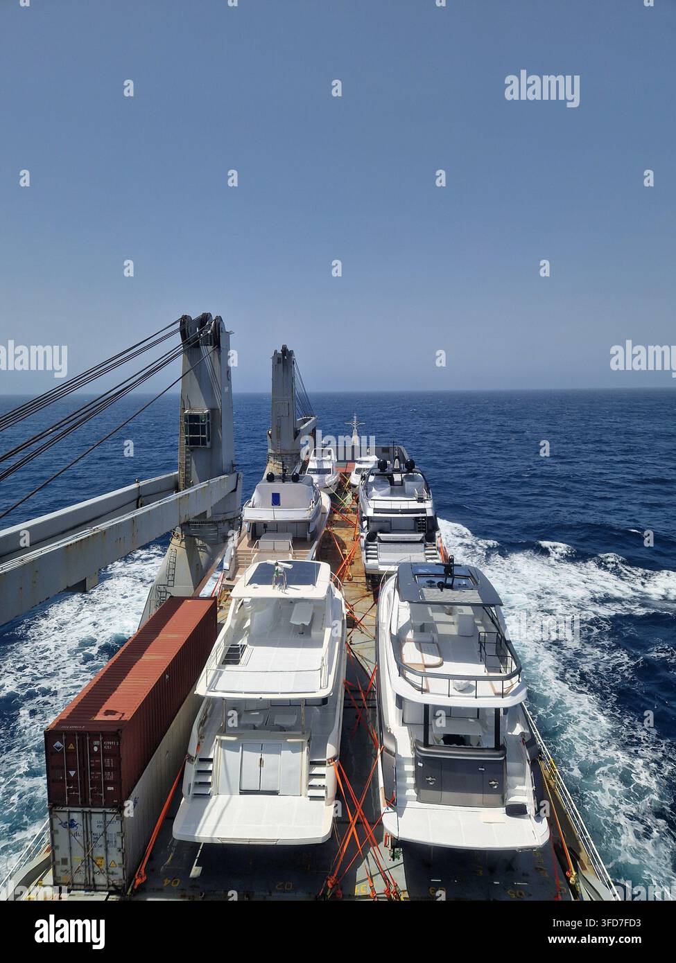 High angle view of a heavy lift cargo ship in open sea carrying  luxury yachts and shipping containers secured on deck under a clear blue sky - Smartphone Captured Stock Image