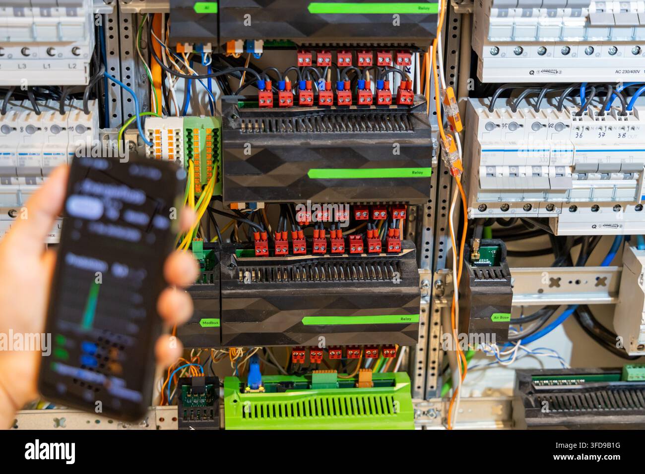 A hand holds a smartphone displaying a control interface next to a complex electrical panel with relays and wiring Stock Photo