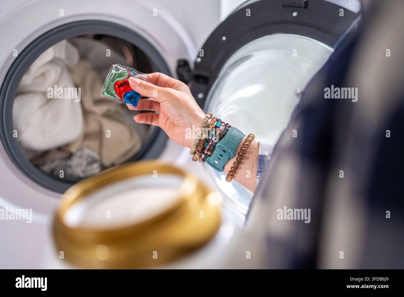 Hand loads colorful laundry detergent pod into open front-loading washing machine. Stock Photo