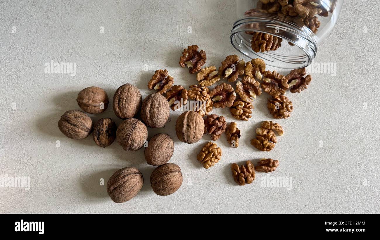 Whole walnuts and walnut kernels arranged as if spilling from a glass jar on a light surface, with generous copy space. Stock Photo