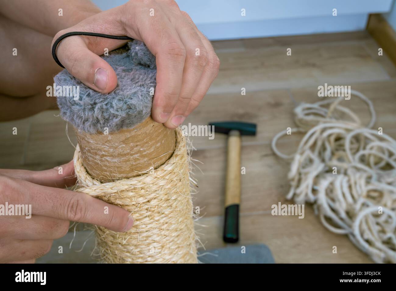 Hands repair a cat scratching post by replacing worn sisal rope around a cardboard tube, with loose cord and hand tools arranged on a wooden workbenc Stock Photo