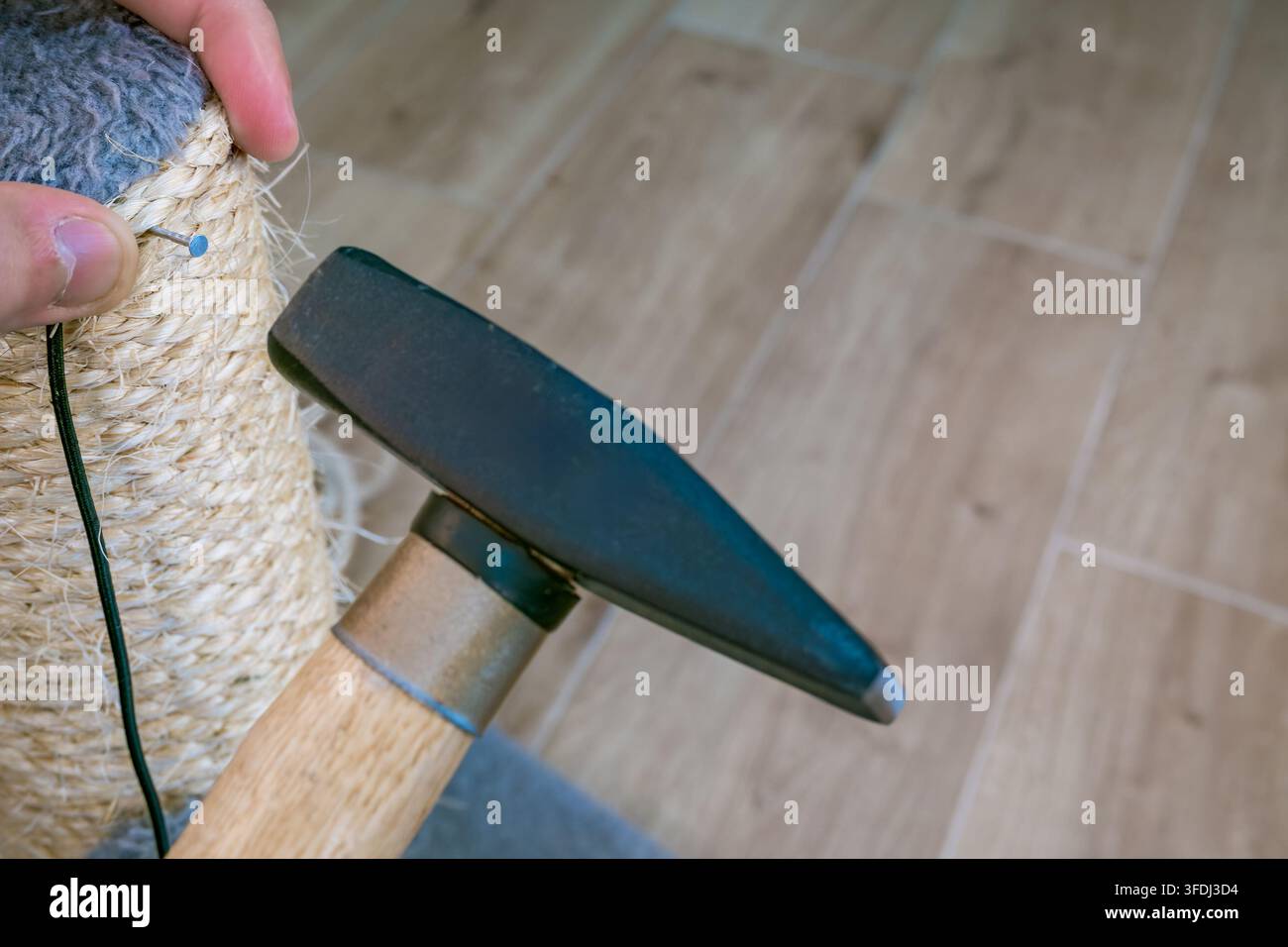Hands secure fresh sisal rope around a worn scratching post while a hammer drives a fastener into the fiber-wrapped surface on a wooden floo Stock Photo