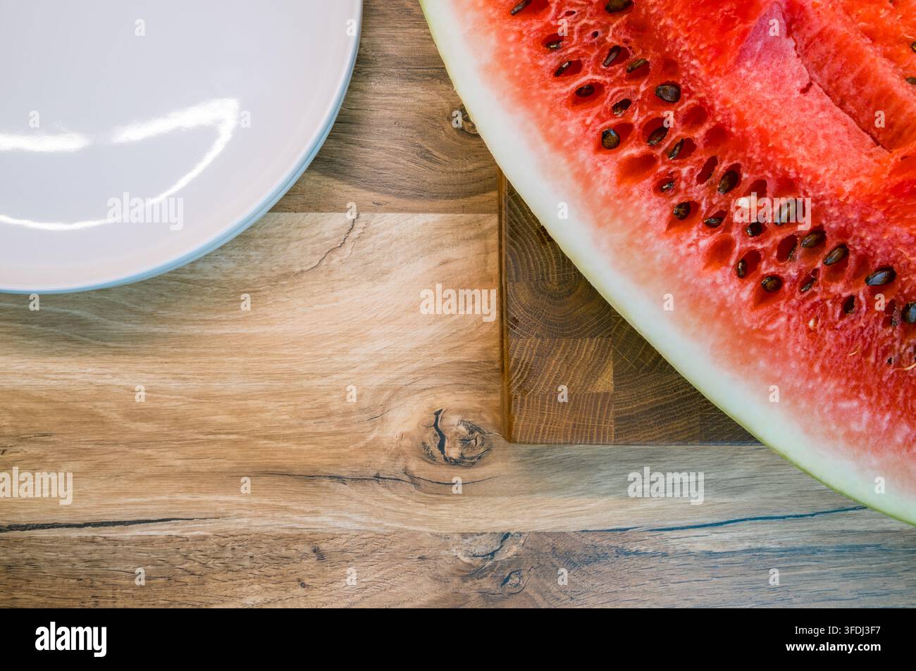 Overhead view of a seeded watermelon wedge on a wooden cutting board beside an empty white plate on a rustic wood tableto Stock Photo