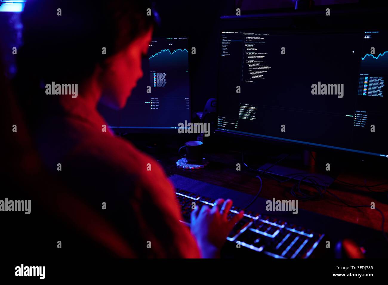 Hacker working late at night, typing on a glowing keyboard with multiple screens displaying code and data Stock Photo