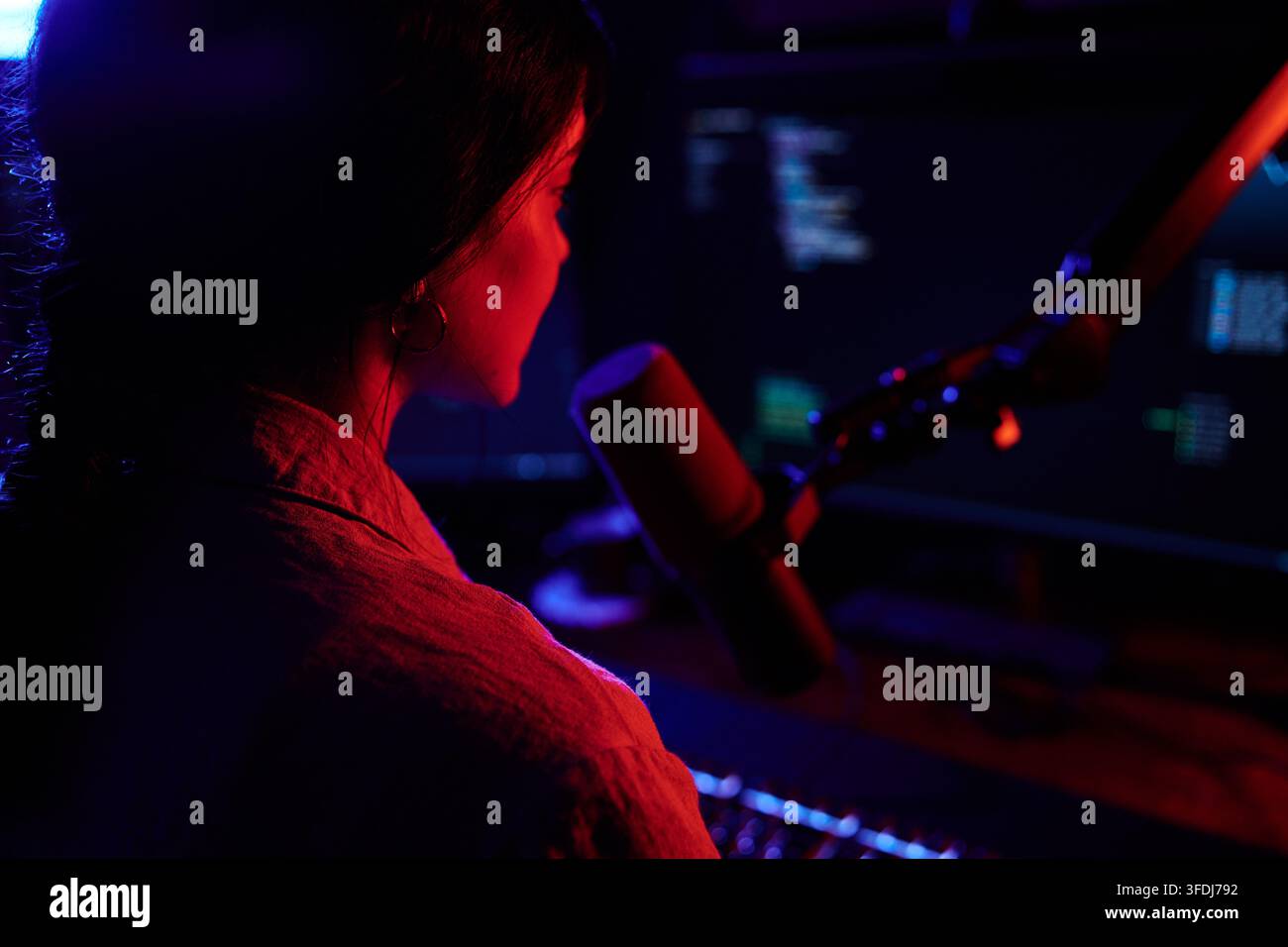 Female programmer focused on screen, working with computer code and microphone, surrounded by neon red and blue light Stock Photo