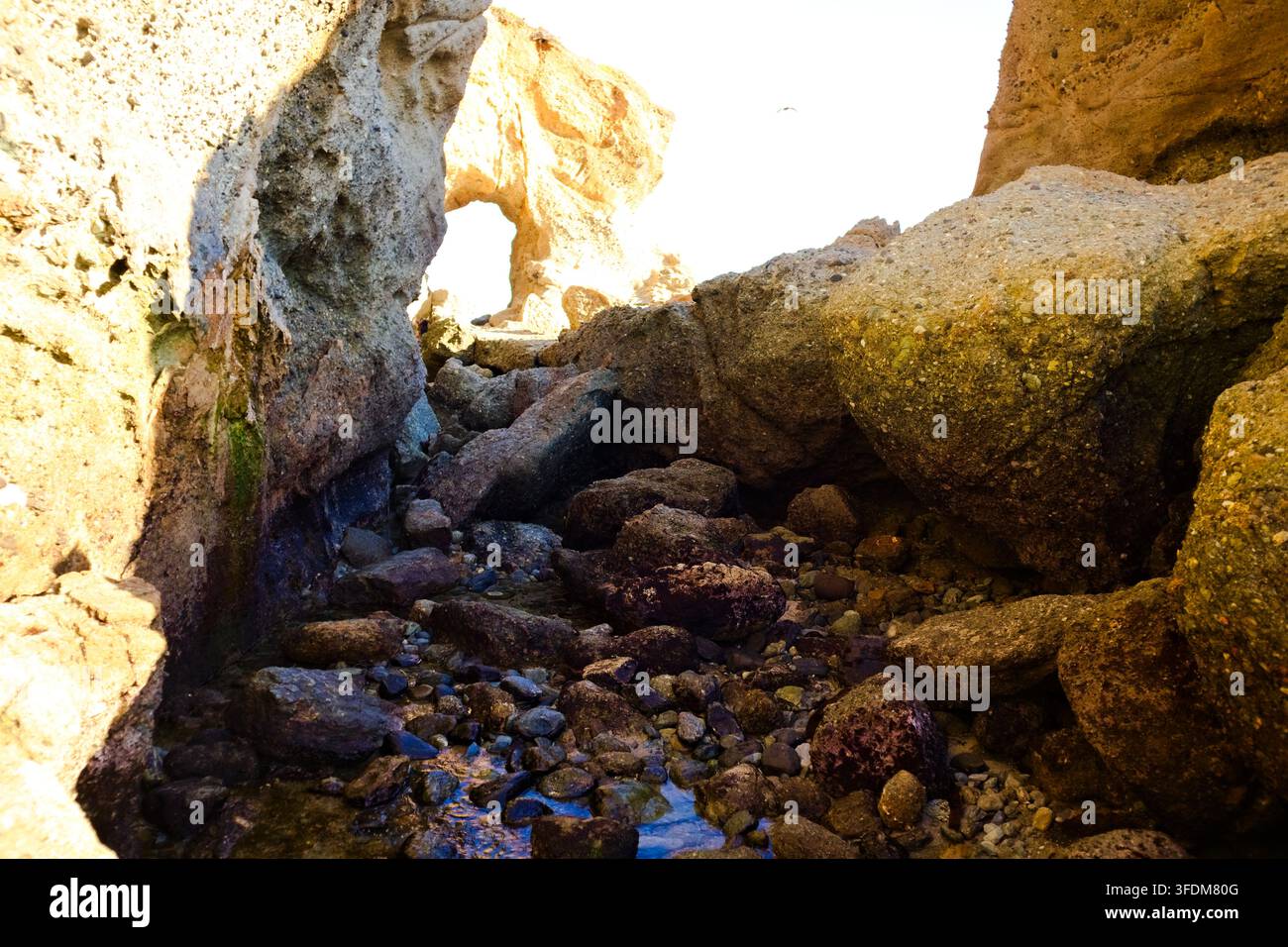 Sunlight shines through a rocky archway over a rugged stream. Stones ...