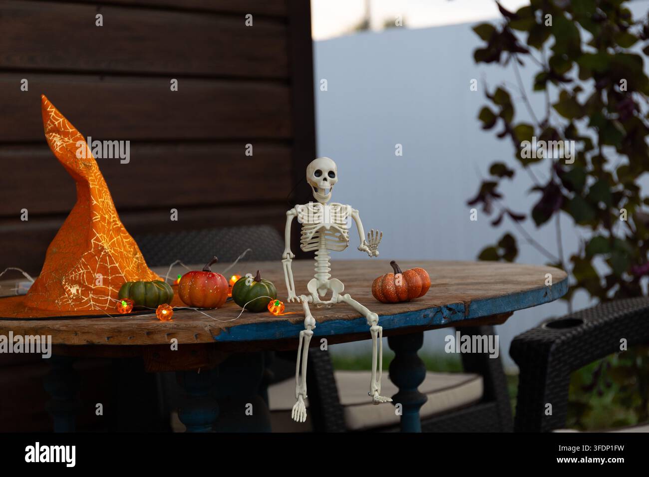 A small plastic Halloween skeleton figurine sits on a wooden veranda table, raises one arm as if waving, surrounded by pumpkins, a witch hat, and stri Stock Photo