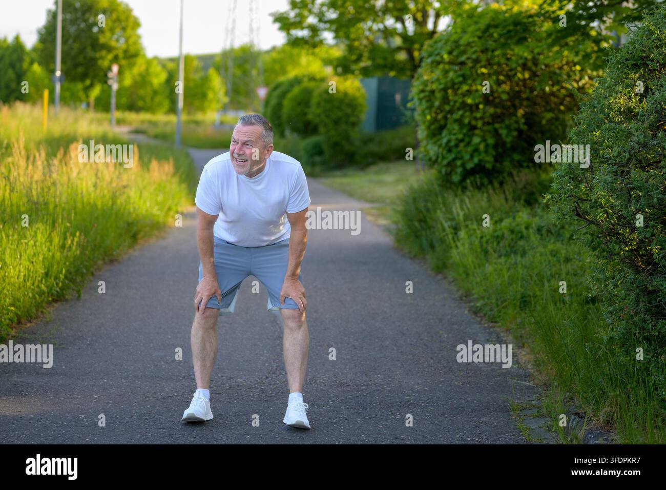 Senior man taking a short recovery break after jogging outdoors on a sunny pathway. Authentic healthy aging, endurance and active lifestyle concept wi Stock Photo