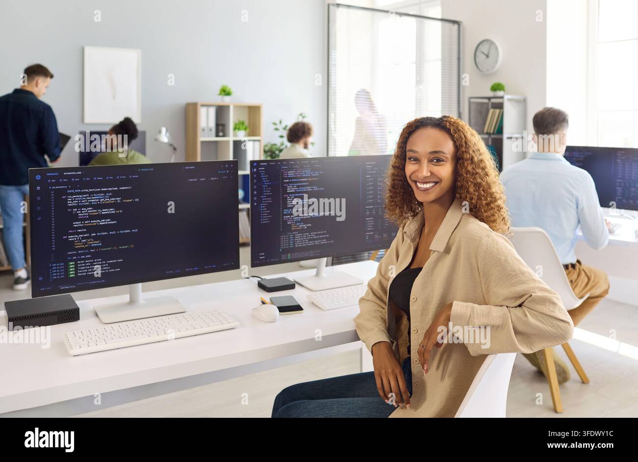 Confident woman programmer at dual monitors in bright collaborative tech office Stock Photo