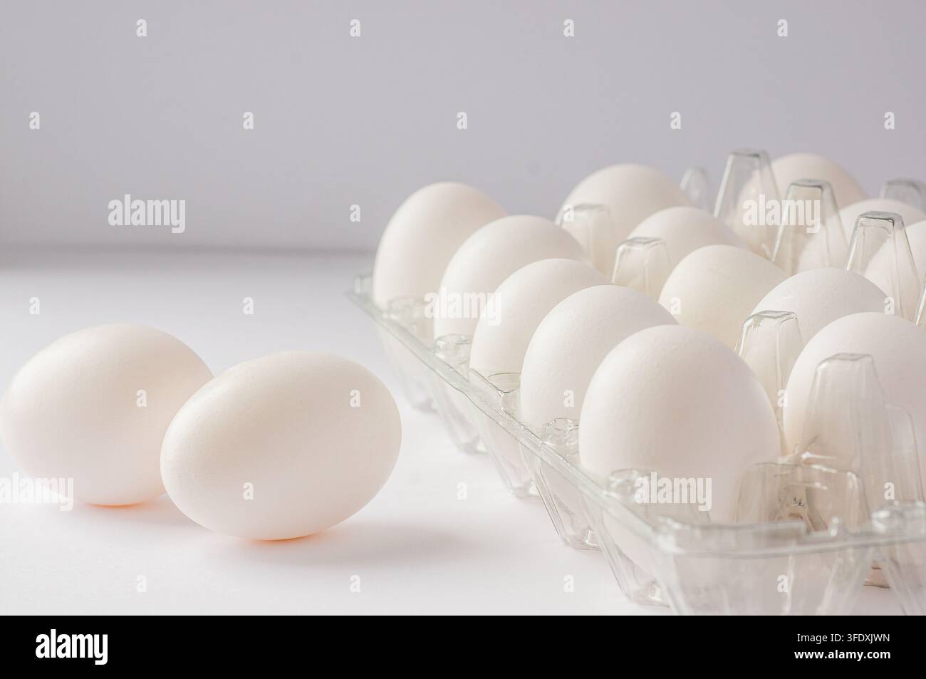 Fresh white eggs in a clear plastic carton with two eggs lying outside on a white surface Stock Photo