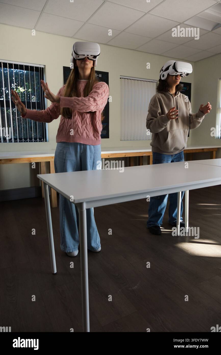 Diverse female youths wearing white VR headsets, standing behind white table in classroom gesturing Stock Photo