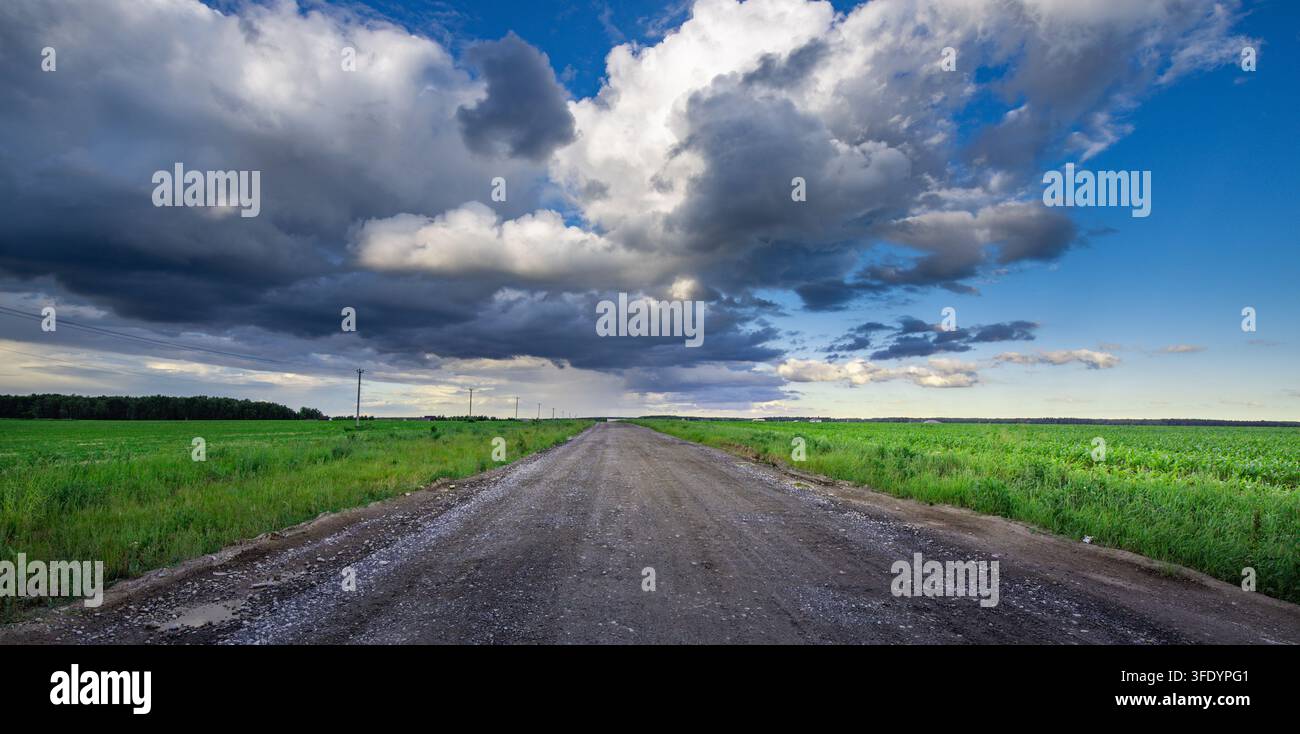 Wide gravel path cuts through vibrant green fields. Sky swirls with dramatic, brooding clouds above Stock Photo