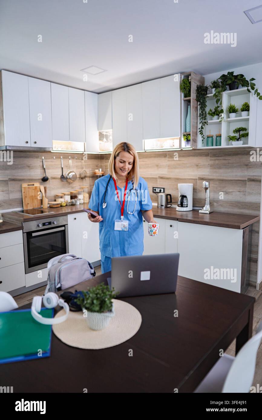 Smiling female doctor in blue uniform drinking coffee and using smartphone and laptop in kitchen Stock Photo