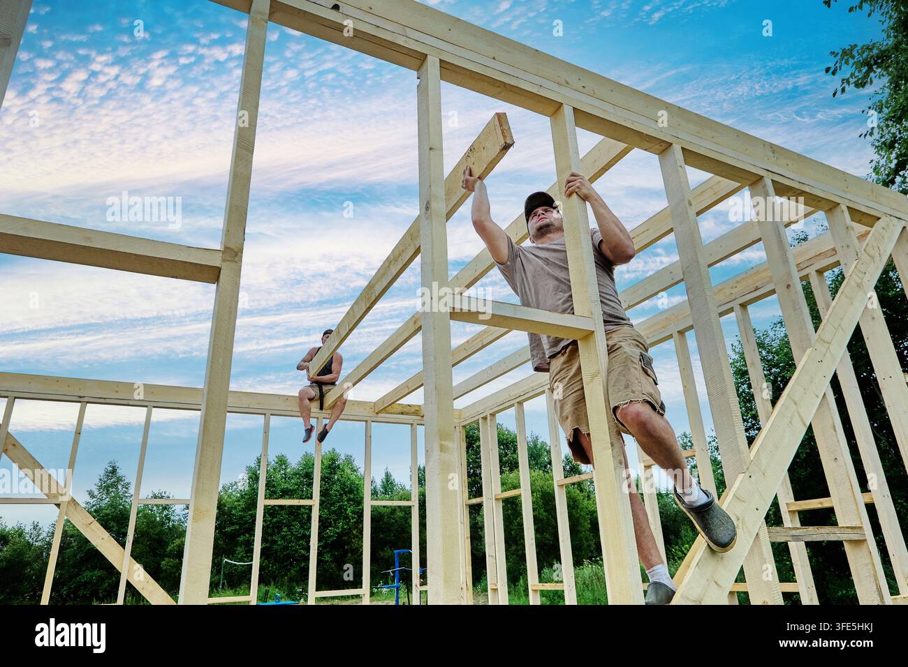 Construction team positions ceiling beams across framework, assembling singlestorey wooden frame house. Stock Photo