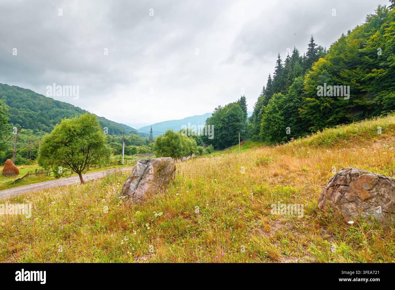 rural landscape. fence near road through field and forest to the high mountains. countryside scenery of ukraine in summer with overcast sky Stock Photo