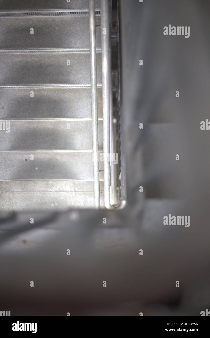 A high-angle view looking down a metal staircase with steel handrails and blurred foreground framing in an industrial building. Stock Photo