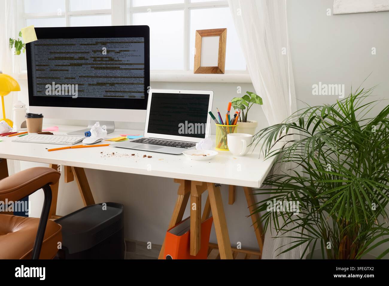 Software development and remote work burnout, messy home office desk with computer and laptop showing programming code in a bright sunlit workspace Stock Photo