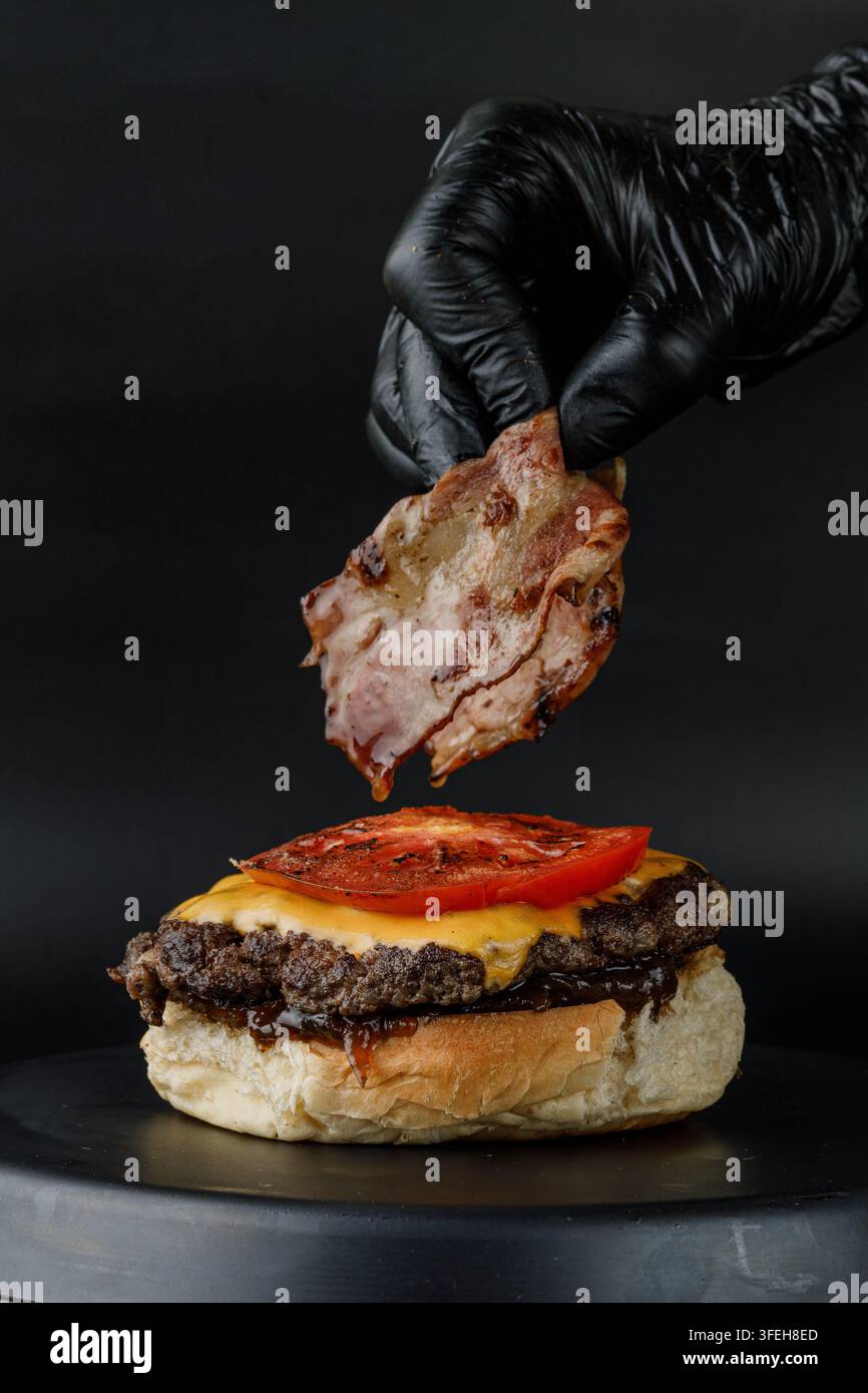 Gloved hand carefully placing bacon on a burger Stock Photo