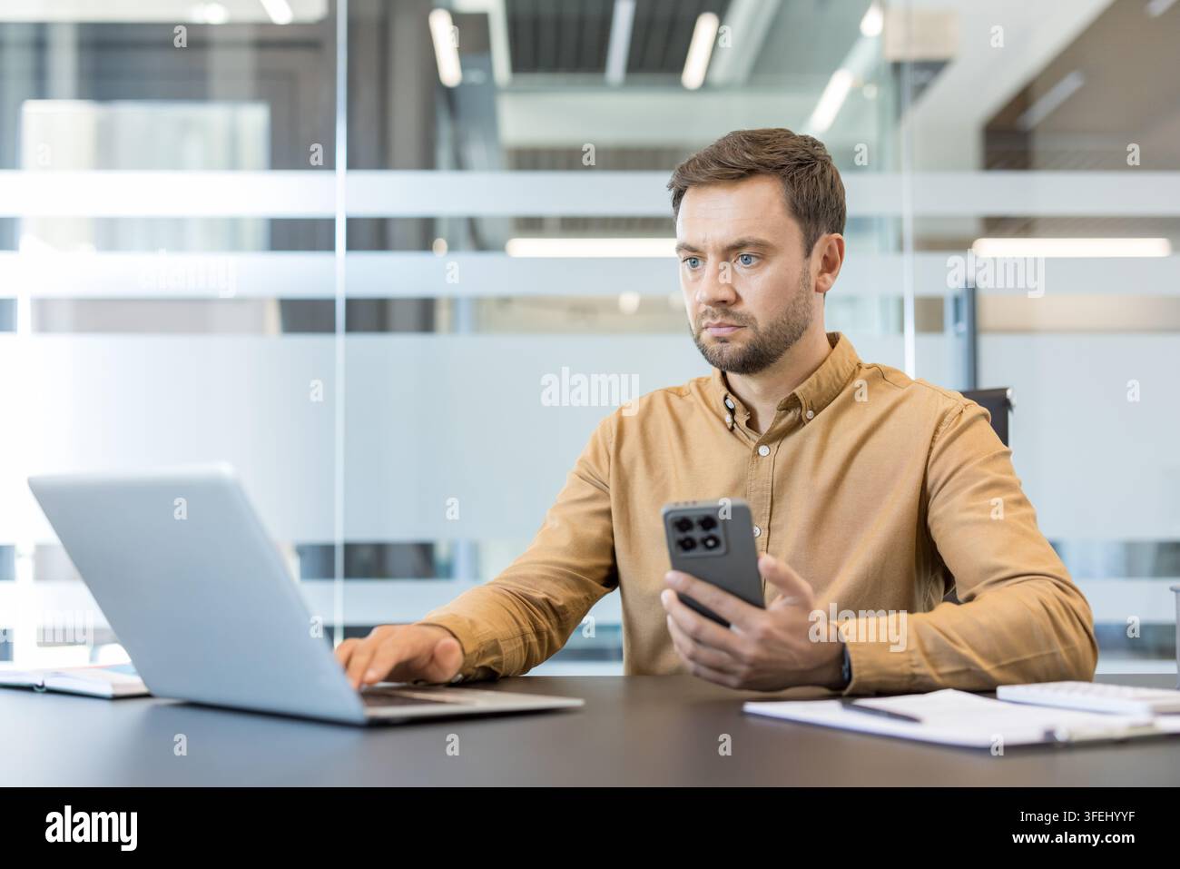 Professional man working on a laptop computer while simultaneously holding and checking a mobile phone, balancing tasks and staying connected in a contemporary business environment Stock Photo