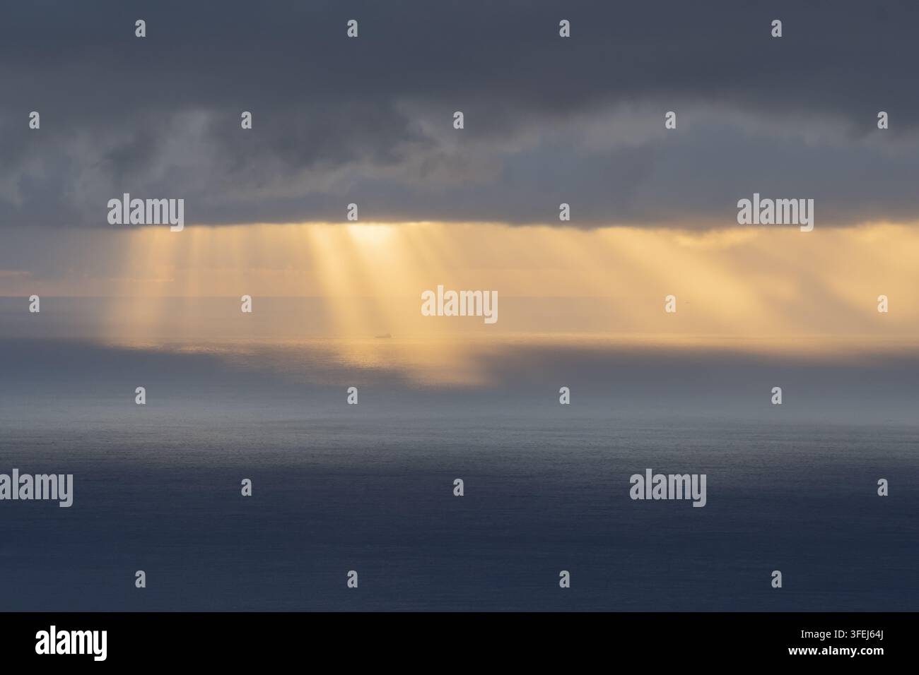 A dramatic dawn over Madeira shows golden sunlight streaming through heavy clouds and reflecting across the calm surface of the Atlantic Ocean. Stock Photo