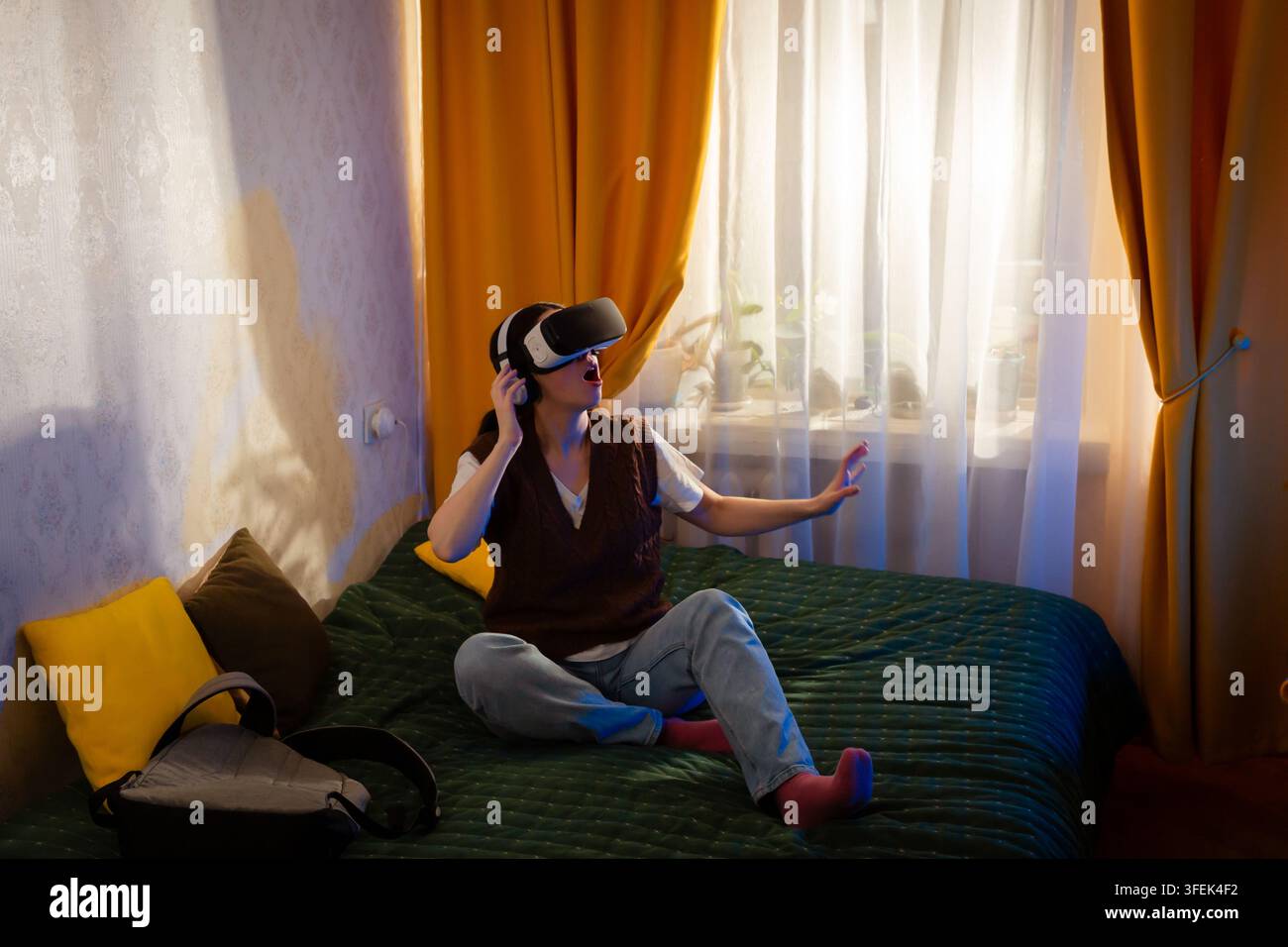 Stylish young woman uses VR glasses in a warm bedroom interior. Digital interaction and future technologies in a home setting. Mid shot. Stock Photo