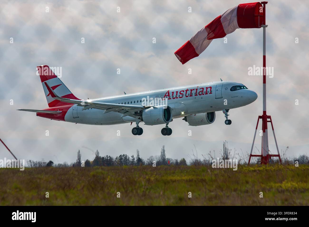 An Austrian Airlines Airbus A220 descends toward the runway with landing gear extended, a red-and-white windsock indicating wind direction. Stock Photo