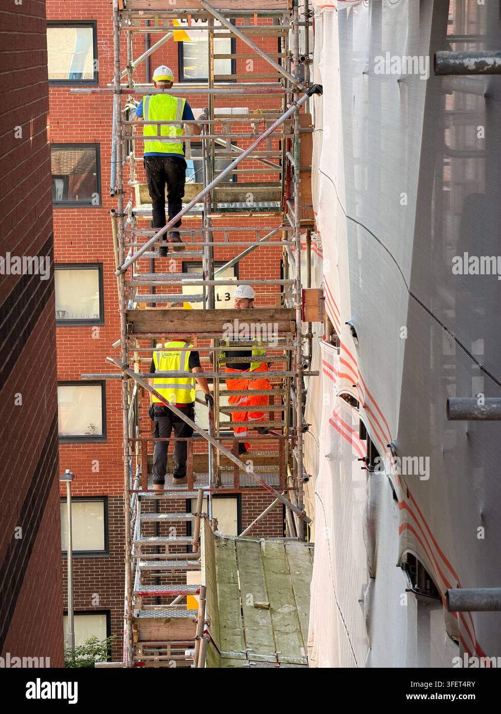 Three workers on multi level scaffolding between brick and sheet covered buildings. Stock Photo