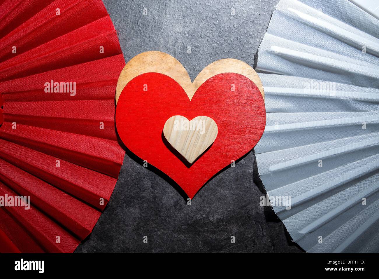 Artistic composition featuring nested heart shapes in red and wood, positioned between decorative red and silver paper fans against a dark textured ba Stock Photo