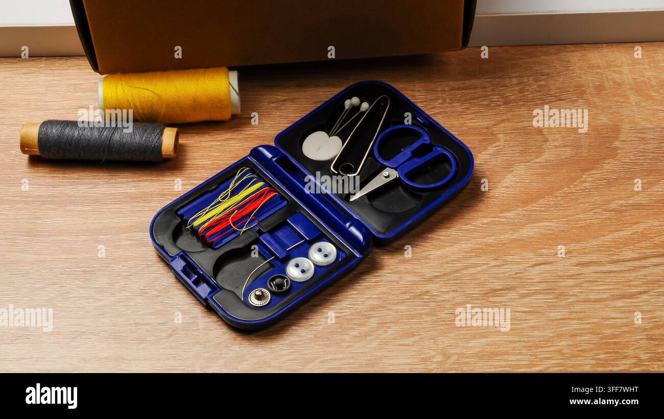 A small, portable sewing kit containing essential mending supplies like colorful threads, needles, a safety pin, and buttons, laid out on a textured w Stock Photo