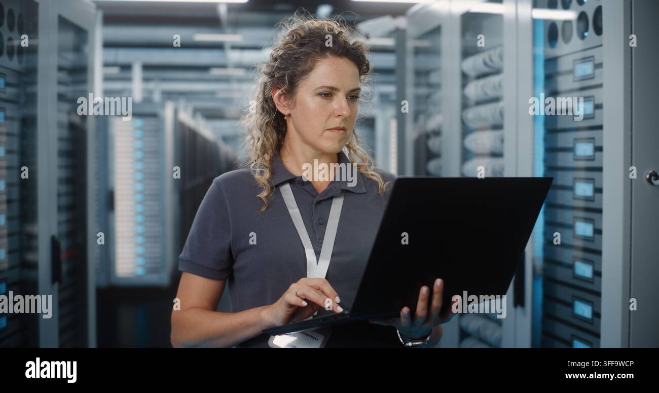 Focused Female Network Engineer Types on Laptop Computer While Standing Near Server Cabinets in Data Center. Hardware Configuration and Server Diagnostics Ensure Continuous Cloud Connectivity. Stock Photo