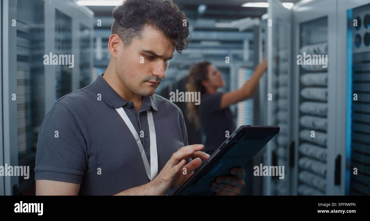 Standing in Server Room, Technician Analyzes Data Metrics on Tablet as Teammate Configures Server Rack Components. Mobile Diagnostic Tools and Physical Maintenance Optimize Cloud Server Efficiency. Stock Photo