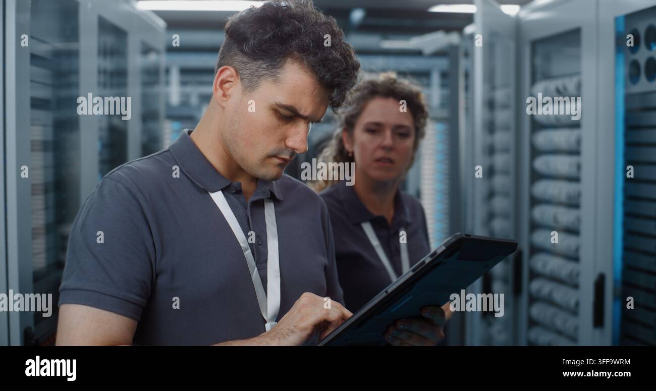 Standing in Server Room, Technician Analyzes Data Metrics on Tablet as Teammate Configures Server Rack Components. Mobile Diagnostic Tools and Physical Maintenance Optimize Cloud Server Efficiency. Stock Photo