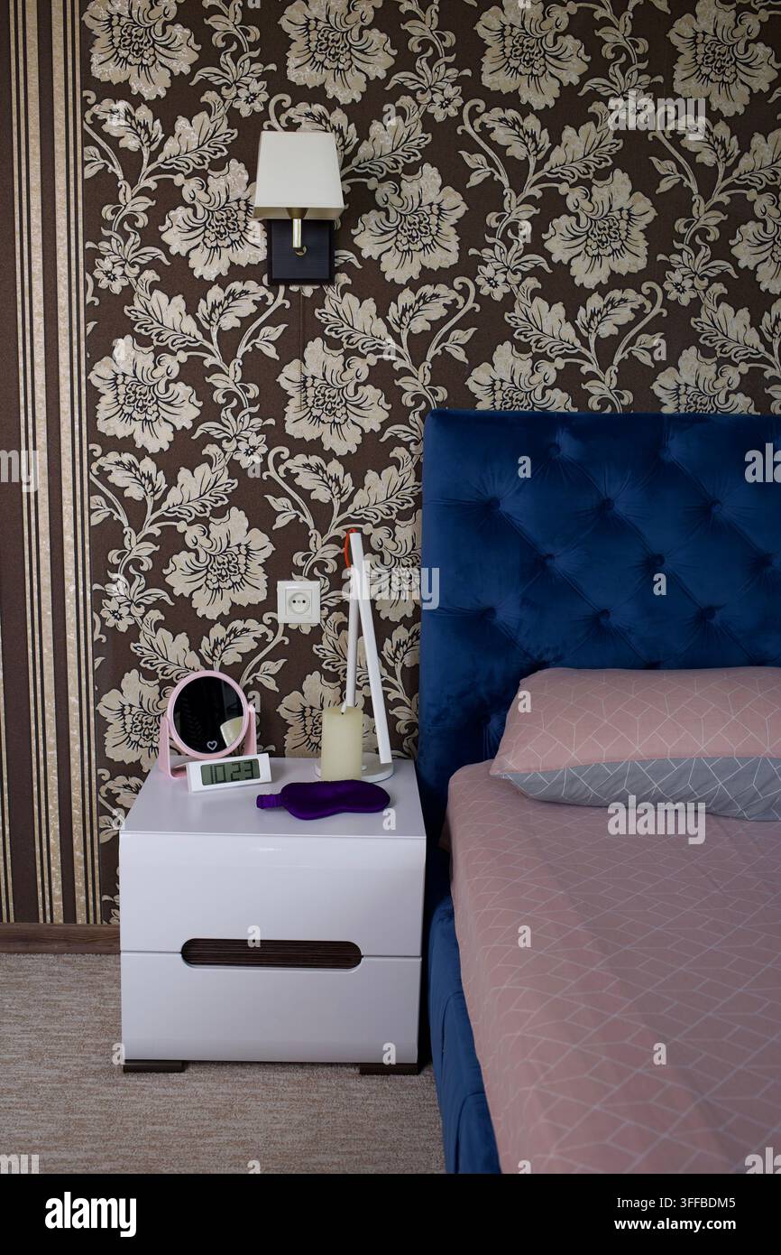 Bedroom bedside scene with patterned beige floral wallpaper, square wall lamp with white shade, deep blue tufted headboard Stock Photo