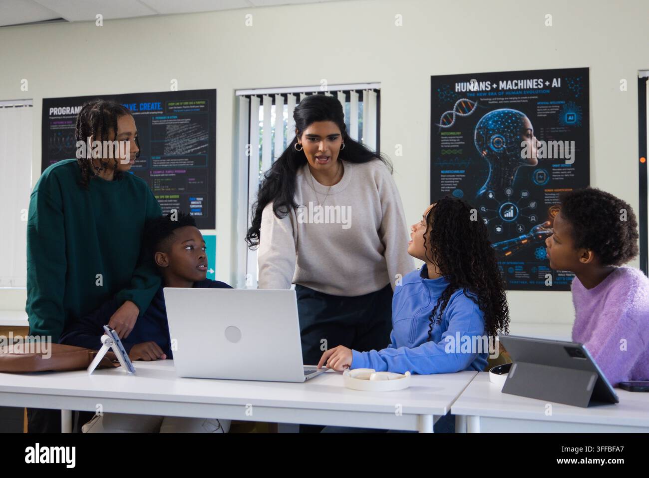 Diverse instructor with youth students leaning over silver laptop, teaching coding in classroom lab Stock Photo