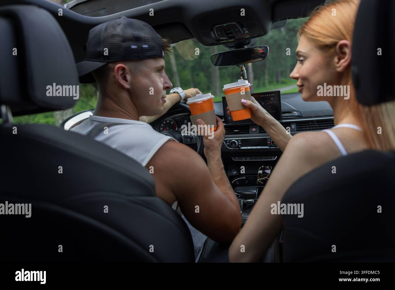 Young couple inside a car holding takeaway coffee cups, looking at each other and smiling during a road trip break. Driver and redhead passenger enjoy Stock Photo