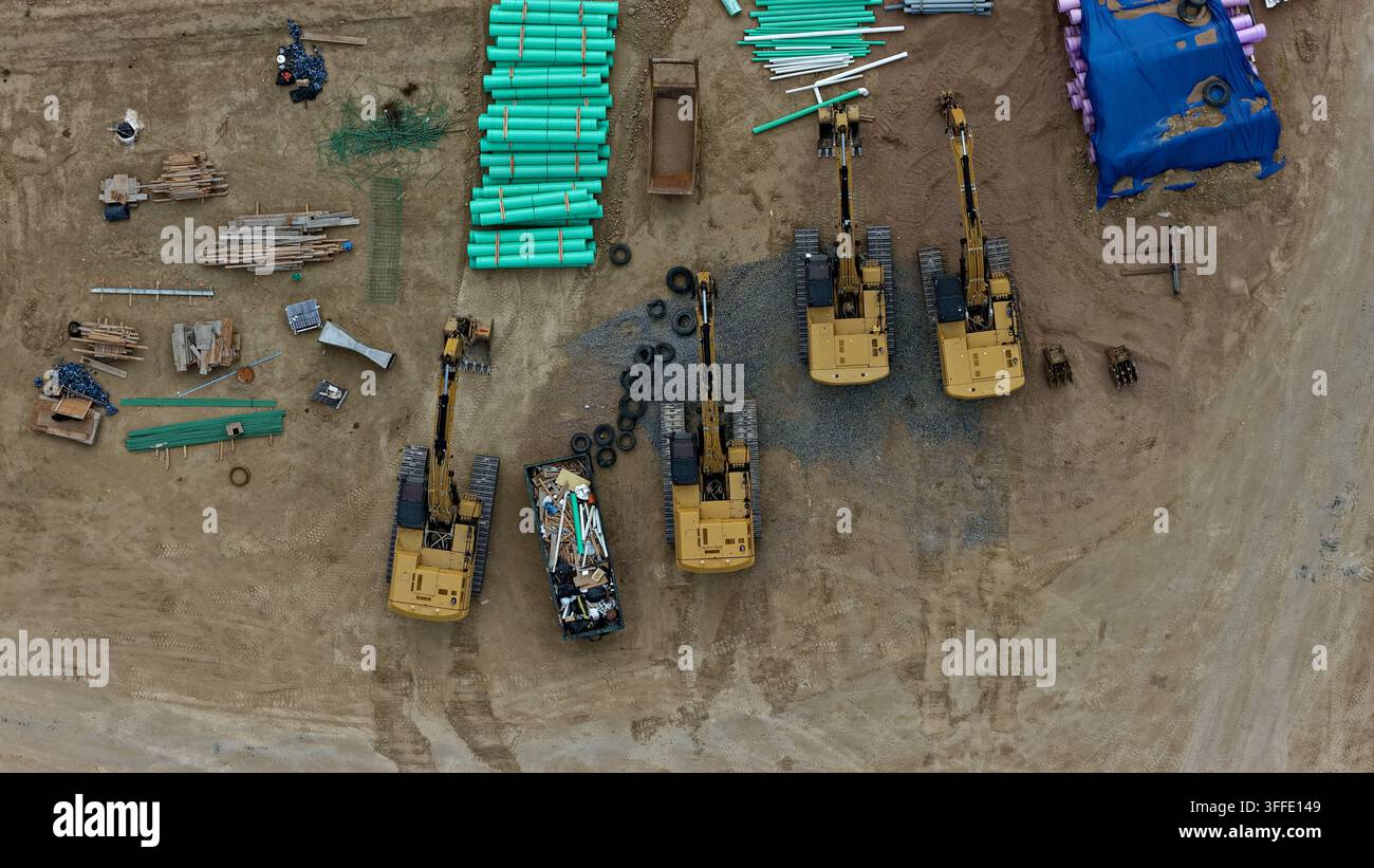 Aerial view of construction site featuring multiple yellow excavators, stacks of green pipes, and various building materials on dirt ground in Longmon Stock Photo