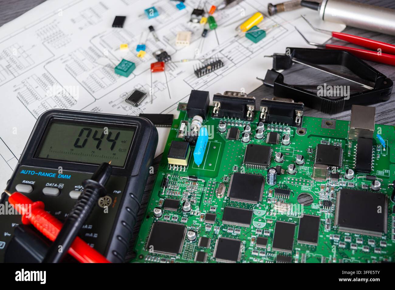 A person tests a circuit board with a multimeter while various electronic components are scattered on the work table. Stock Photo